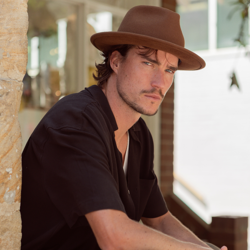 Male Model sitting in front of sandstone wall, wearing a brown felt hat