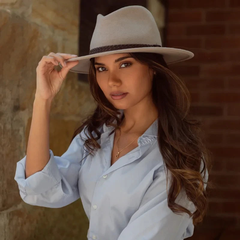 Female Model wearing a sand coloured Felt Hat sitting in front of a stone wall and lightly touching her hat